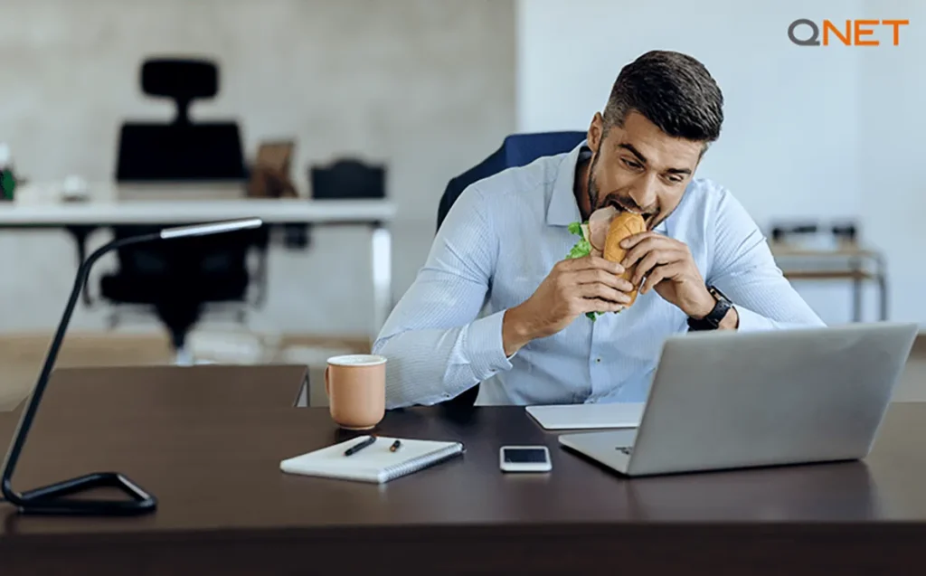 A young man following an unhealthy lifestyle, eating a burger while working in his office