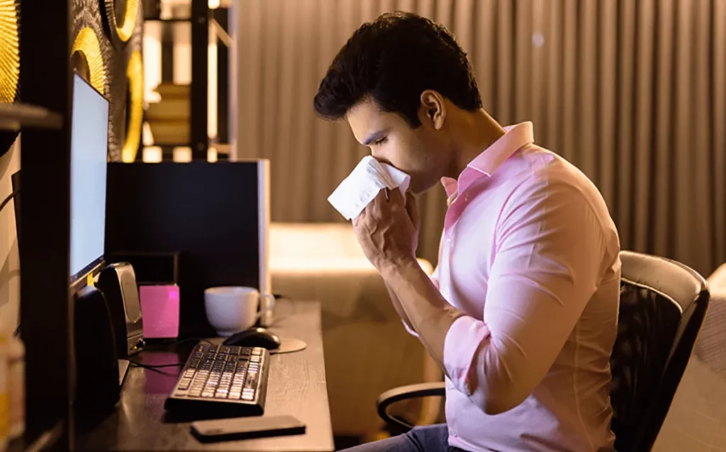 Indian man sneezing into a white cloth while working in front of a computer due to allergy.